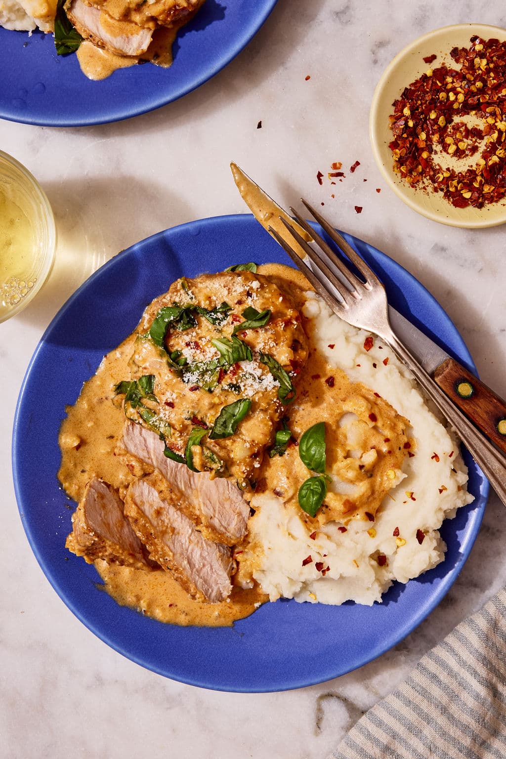 Overhead view of sliced Marry Me Pork Chops covered in a creamy sun-dried tomato Parmesan sauce with fresh basil, served alongside mashed potatoes on a blue plate with a fork and knife and a glass of white wine nearby.