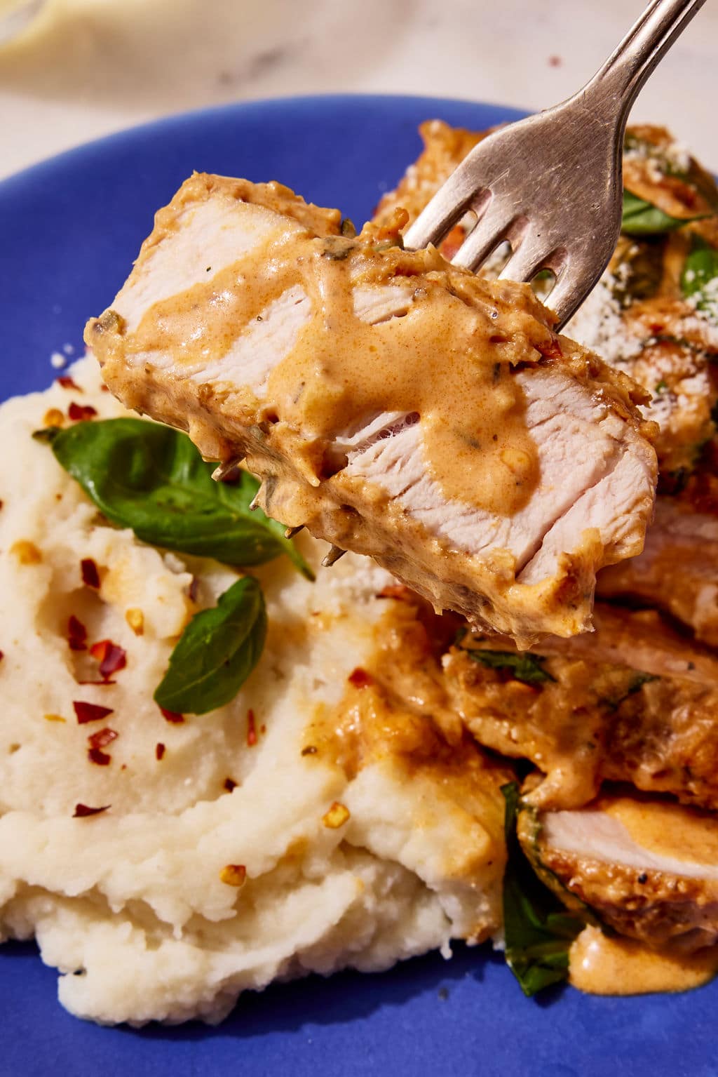Close-up of a fork holding a tender slice of Marry Me Pork Chop coated in creamy sun-dried tomato Parmesan sauce, with mashed potatoes, fresh basil, and additional sliced pork chops on a blue plate in the background.