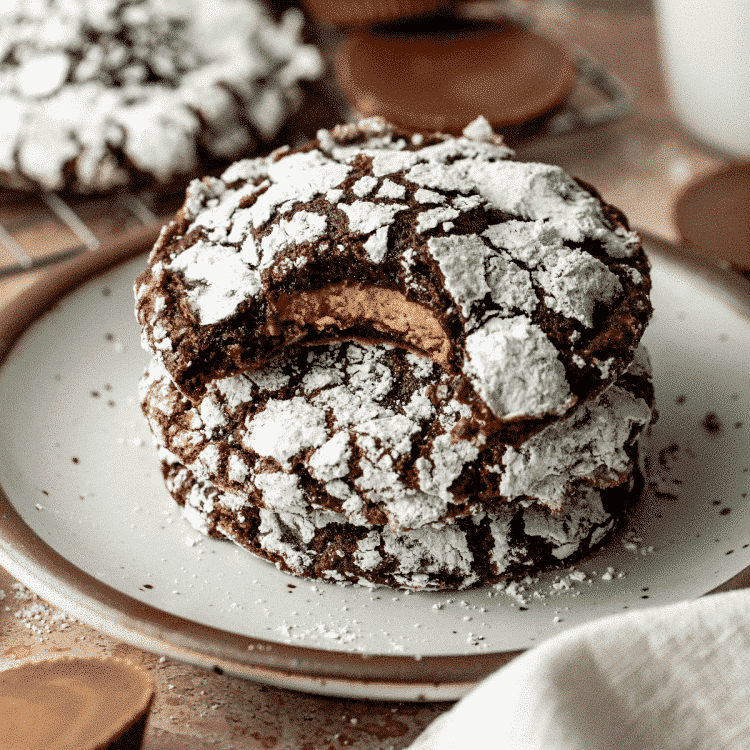 Three reese's crinkle cookies stacked on a plate with the top cookie showing the inside of the reese's cup stuffed inside.